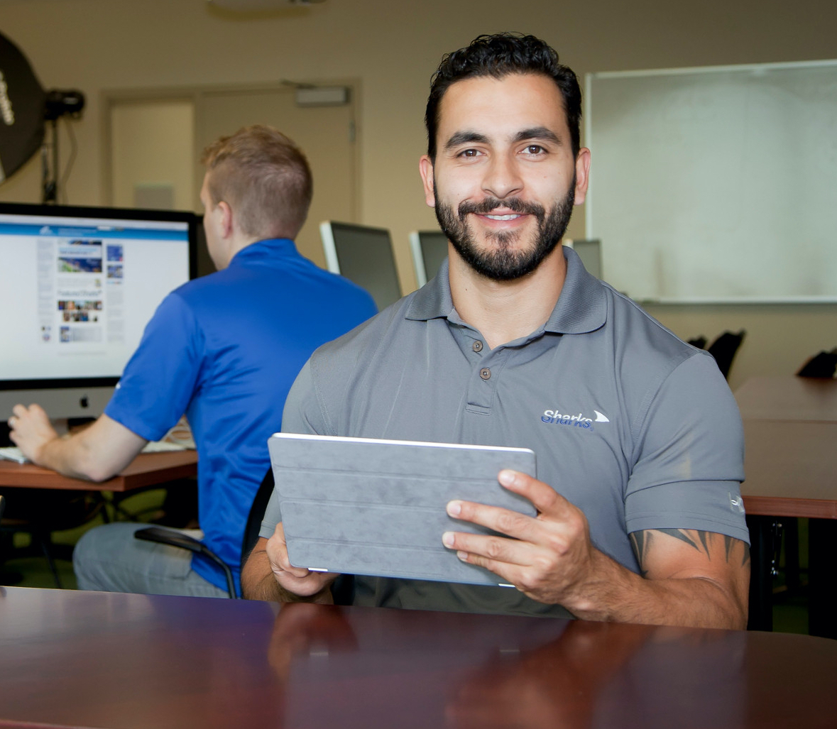 man using a tablet device with another man in the background on a computer